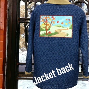 May include: A blue denim jacket with a quilted pattern and a brown corduroy collar. The back of the jacket features an embroidered patch depicting a fall scene with a tree, leaves, and a winding path.