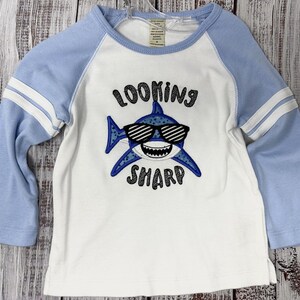 May include: White and light blue raglan-style long-sleeve shirt. The shirt features a shark graphic wearing sunglasses with the text "LOOKING SHARP" in black lettering. The sleeves have white stripes.