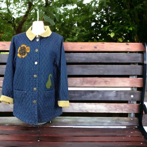 May include: A blue denim jacket with a quilted design and a corduroy collar. The jacket has two patch pockets on the front and features a sunflower and a pear design.
