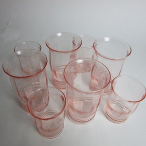 May include: A set of eight pink-hued glass tumblers. The glasses feature a flared rim and a ribbed, faceted design. The glasses are arranged in a circular pattern, highlighting their vintage style. The glassware is suitable for serving drinks.