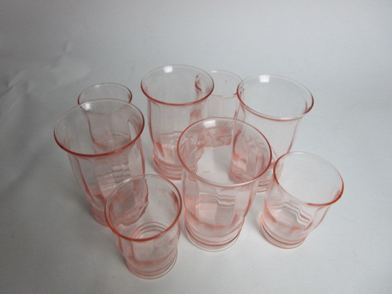 May include: A set of eight pink-hued glass tumblers. The glasses feature a flared rim and a ribbed, faceted design. The glasses are arranged in a circular pattern, highlighting their vintage style. The glassware is suitable for serving drinks.
