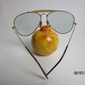 May include: Gold aviator sunglasses with brown temples and gray lenses. The sunglasses are resting on a yellow and orange fruit.