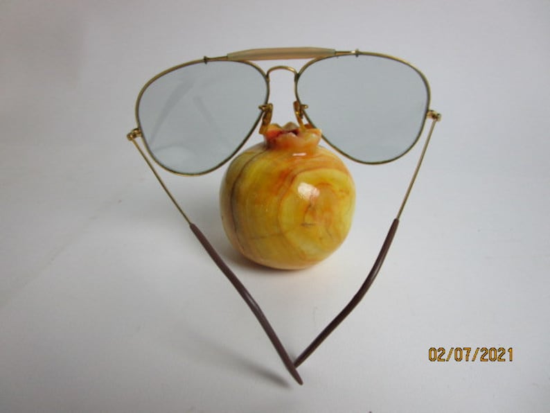 May include: Gold aviator sunglasses with brown temples and gray lenses. The sunglasses are resting on a yellow and orange fruit.