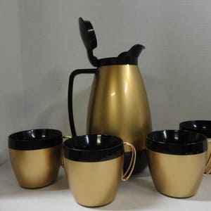 May include: A gold-colored insulated carafe with a black lid and handle, accompanied by four matching gold and black mugs. The set is designed for serving hot beverages and has a retro aesthetic.