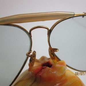 May include: Gold-tone aviator sunglasses with a brown plastic nose bridge and blue tinted lenses. The sunglasses are resting on a light brown surface.