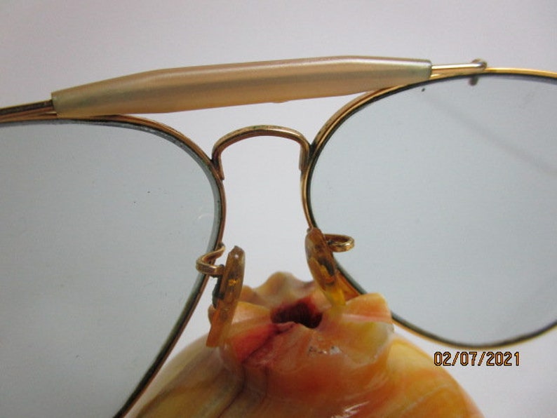 May include: Gold-tone aviator sunglasses with a brown plastic nose bridge and blue tinted lenses. The sunglasses are resting on a light brown surface.