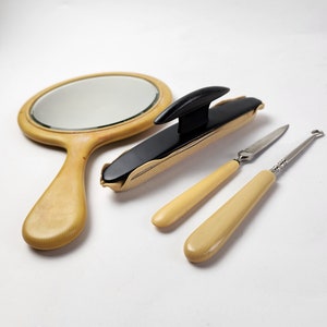 May include: A vintage vanity set featuring a hand mirror, a black and gold shoe horn, and two ivory-colored handled tools. The mirror has a light wood frame. The tools include a knife and a hook, all set against a white background.