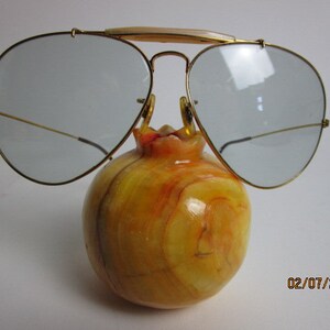 May include: Gold aviator sunglasses with clear lenses resting on a yellow and orange marble pomegranate.