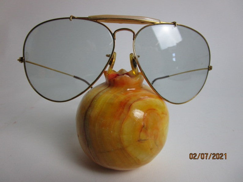 May include: Gold aviator sunglasses with clear lenses resting on a yellow and orange marble pomegranate.