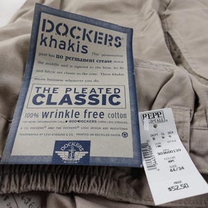 May include: A beige pair of Dockers khakis with a tag attached. The tag is white with blue text and a Dockers logo. The tag says "Dockers khakis, The Pleated Classic, 100% wrinkle free cotton". The tag also includes a phone number and information about the company. The tag is attached to a second tag that is white with black text and says "Pepp & Tall, Dept. CL Sea 36 16, VDR 11, Style 303600139, Color Rope, Size 44/34, Price $52.50".