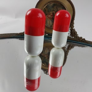 May include: Two decorative pill-shaped objects, each with a red top and a white bottom. The objects are reflected in a mirror. A decorative, ornate frame is in the background. The objects are likely bottle stoppers or decorative items.