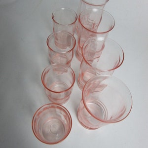 May include: A set of seven vintage pink glass tumblers. The glasses are in various sizes and shapes, with a delicate, translucent pink colour. The arrangement highlights the different heights and designs of the glassware.