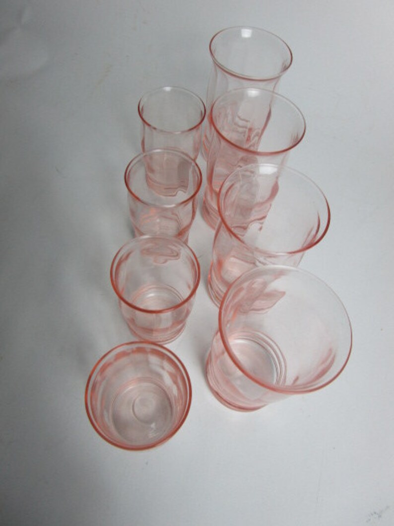 May include: A set of seven vintage pink glass tumblers. The glasses are in various sizes and shapes, with a delicate, translucent pink colour. The arrangement highlights the different heights and designs of the glassware.