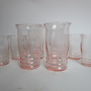 May include: A collection of seven pink-hued glass tumblers. The glasses feature a flared rim and a ribbed design on the lower section. The glasses are displayed on a white surface.
