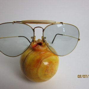 May include: Gold aviator sunglasses with a light blue lens and a white plastic nose bridge. The sunglasses are resting on a yellow and orange marble object.