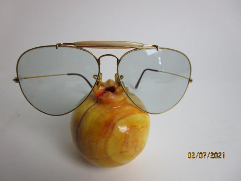 May include: Gold aviator sunglasses with a light blue lens and a white plastic nose bridge. The sunglasses are resting on a yellow and orange marble object.