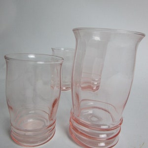May include: Three pink glass tumblers. The glasses have a rounded shape with a flared top and a ribbed base. The transparent glass allows light to pass through. The pink hue gives them a vintage feel. The tumblers are of varying sizes.