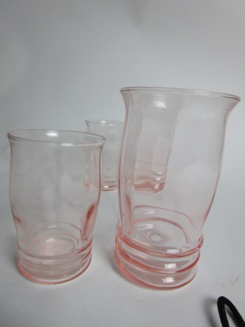 May include: Three pink glass tumblers. The glasses have a rounded shape with a flared top and a ribbed base. The transparent glass allows light to pass through. The pink hue gives them a vintage feel. The tumblers are of varying sizes.