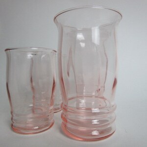 May include: Three vintage pink glass tumblers. The glasses have a flared rim and a ribbed base. The tallest glass is approximately 18 cm tall. The other two glasses are shorter and have a similar design. The glasses are made of a light pink glass.