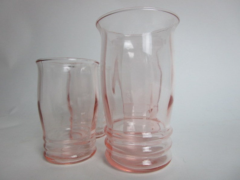 May include: Three vintage pink glass tumblers. The glasses have a flared rim and a ribbed base. The tallest glass is approximately 18 cm tall. The other two glasses are shorter and have a similar design. The glasses are made of a light pink glass.