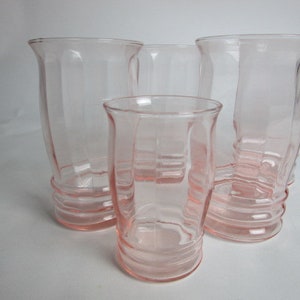 May include: Four vintage pink glass vases of varying sizes. The vases have a ribbed design and flared rims. The glass is a soft, translucent pink. The tallest vase is approximately 18 cm tall. The vases are arranged on a white surface.
