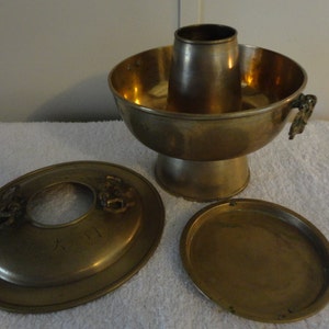 May include: A brass hot pot with a central chimney, a bowl-shaped body, and a pedestal base. It includes a lid with dragon head handles and a small, oval-shaped plate. The hot pot is designed for cooking and serving food.