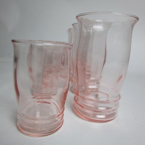 May include: Four pink glass tumblers are shown. The glasses have a curved shape and a ribbed base. The glass is a soft pink colour, and the tumblers are arranged on a white surface.