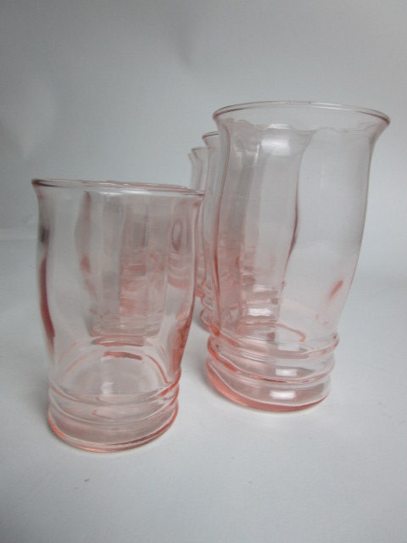 May include: Four pink glass tumblers are shown. The glasses have a curved shape and a ribbed base. The glass is a soft pink colour, and the tumblers are arranged on a white surface.