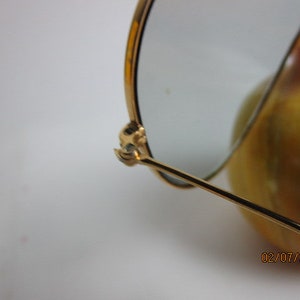 May include: Gold-toned aviator sunglasses with a broken arm. The sunglasses are resting on a yellow and orange surface.