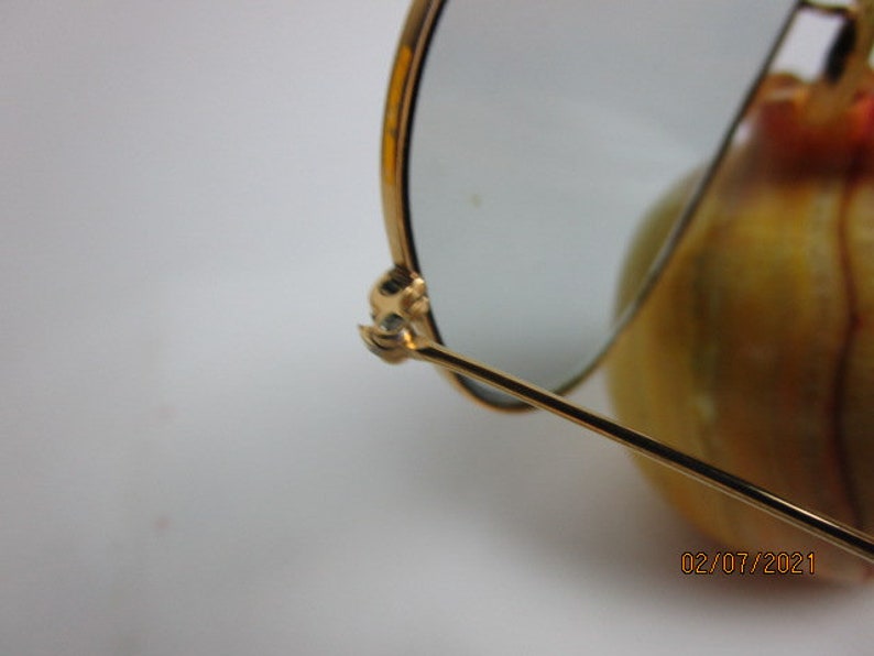 May include: Gold-toned aviator sunglasses with a broken arm. The sunglasses are resting on a yellow and orange surface.