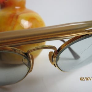 May include: Gold-tone metal glasses with a tortoise shell nose bridge and clear lenses. The lenses have a small crack in the upper right corner. The glasses are resting on a yellow and orange marble surface.