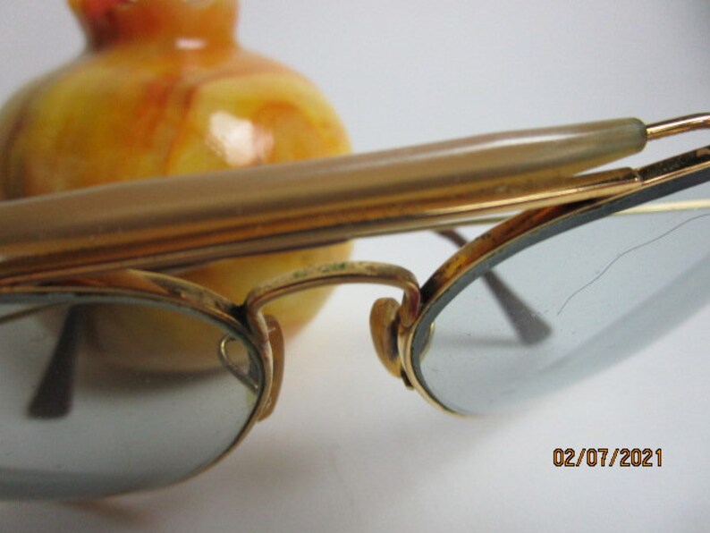 May include: Gold-tone metal glasses with a tortoise shell nose bridge and clear lenses. The lenses have a small crack in the upper right corner. The glasses are resting on a yellow and orange marble surface.