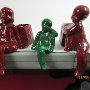 May include: Three ceramic figurines in a row. Two are a deep red color, and one is green. Each figurine is posed as if sitting with their legs dangling. The red figurines are holding a small, textured, red container. The green figurine is leaning on its arm.
