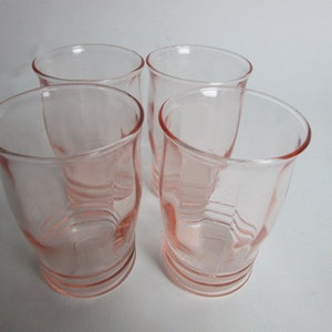 May include: Four vintage pink glass tumblers. The glasses have a rounded shape with a slightly flared rim and a ridged base. The pink colour is consistent throughout the glass, giving them a delicate appearance. The glasses are arranged on a white surface.