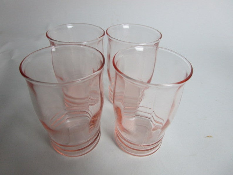 May include: Four vintage pink glass tumblers. The glasses have a rounded shape with a slightly flared rim and a ridged base. The pink colour is consistent throughout the glass, giving them a delicate appearance. The glasses are arranged on a white surface.