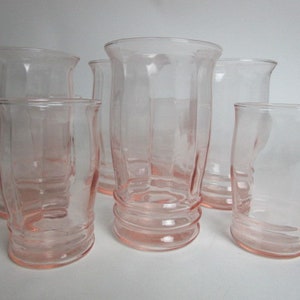 May include: A collection of six vintage pink glass tumblers. The glasses feature a curved, ribbed design at the base and a flared rim. The glasses are empty and are on a white surface.