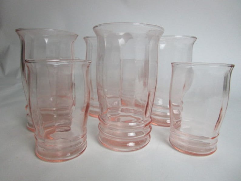 May include: A collection of six vintage pink glass tumblers. The glasses feature a curved, ribbed design at the base and a flared rim. The glasses are empty and are on a white surface.