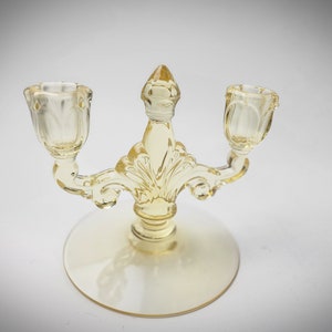 May include: A clear, yellow glass candelabra with two candle holders. The candelabra has a decorative, ornate design with a round base and a central finial. The candle holders are tulip-shaped.