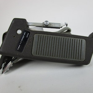 May include: A dark gray Canon C-8 Trigger Grip, made in Japan. The grip features a textured, ridged pad, a silver metal trigger, and a gray strap. The product has a small, rectangular label with the Canon C-8 Trigger Grip text.