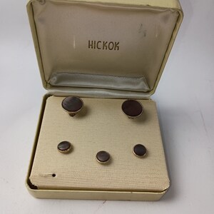 May include: Vintage HICKOK cufflink and shirt stud set in a cream-colored box. The set includes two cufflinks and four shirt studs with dark, iridescent centers and gold-toned rims. The box is open, revealing the contents.