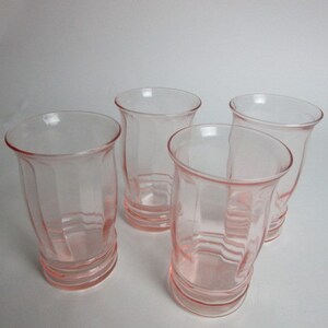 May include: Four vintage pink glass tumblers. Each glass has a flared rim and a ribbed, faceted design. The glasses are a soft pink colour and appear to be made of pressed glass. The glasses are empty and sit on a white surface.