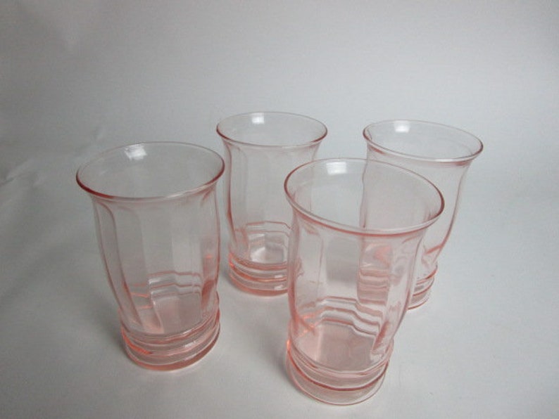 May include: Four vintage pink glass tumblers. Each glass has a flared rim and a ribbed, faceted design. The glasses are a soft pink colour and appear to be made of pressed glass. The glasses are empty and sit on a white surface.