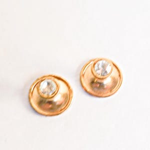 Könnte beinhalten: Ein Paar goldfarbene Ohrstecker. Jeder Ohrring hat einen klaren, runden Edelstein, der in der Mitte eines kreisförmigen, leicht erhabenen Designs gefasst ist. Die Ohrringe werden vor einem weißen Hintergrund präsentiert.