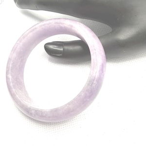 May include: A smooth, polished lavender-colored bangle bracelet with a rounded profile and a translucent, marbled appearance. The circular jewelry piece features subtle variations in light purple tones throughout its solid, continuous band structure.
