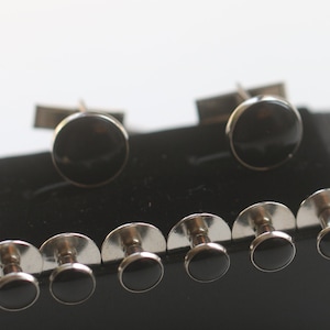 May include: A set of formalwear accessories, including cufflinks and shirt studs. The cufflinks and studs feature a silver-tone metal setting with a round, black, glossy center. The set is displayed on a black velvet surface.