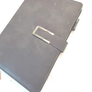 May include: A dark gray rectangular notebook with a smooth matte finish. The cover features a horizontal strap closure secured by a rectangular silver-toned metal buckle. The edges of the notebook are stitched with matching dark gray thread.