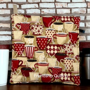 May include: A red and cream quilted hot pad with a coffee cup pattern. The hot pad features a variety of coffee cups with different designs, including stars, stripes, and hearts. The text "Friendship Quilting" is printed on the bottom of the hot pad.