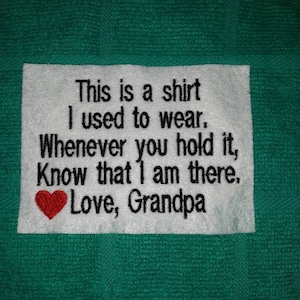 May include: A white rectangular patch embroidered with black text and a red heart, reading "This is a shirt I used to wear. Whenever you hold it, Know that I am there. Love, Grandpa." The patch is on a green fabric.