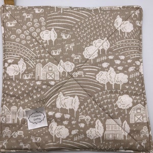 May include: A brown and white quilted hot pad with a farmhouse theme. The pad features a white farmhouse with a red roof, white trees, and white cows. The pad has a brown fabric loop for hanging. The pad has a white label that says "Friendship Quilting".