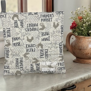 May include: A beige pot holder with a farmhouse theme. It features a repeating pattern of roosters, vines, and the text "Farm Fresh Choice", "Farmer's Market", and "Friendship Quilting".